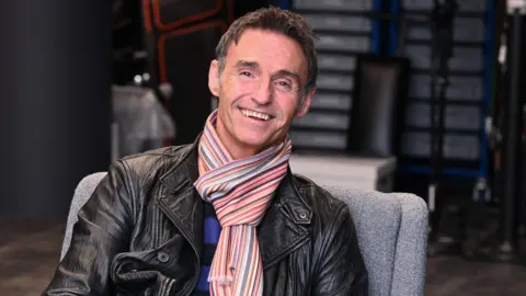 BBC Marti Pellow sits in a grey chair smiling at the camera in the green room at BBC Radio 2. He wears a black leather jacket, a blue and black striped sweater and a multicoloured stripy scarf. A chair and a table are in the background.