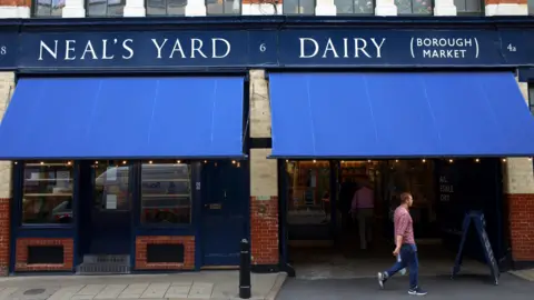 Getty Images The outside of the store in Borough Market, has Neal's Yard Dairy written in white and blue sign with empty shelves in the front window and a man wearing a red chequed shirt and blue jeans walking past the right hand side of the store