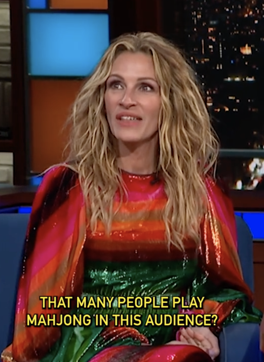 Woman with wavy hair, in a vibrant striped dress, sitting during a talk show, caption reads, "That many people play Mahjong in this audience?"