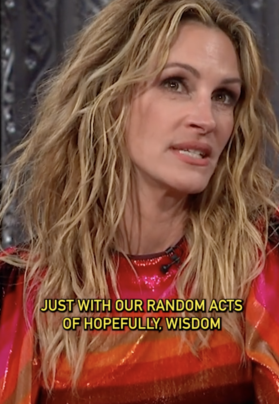 A person with wavy hair in a colorful top is speaking. Subtitles read: "Just with our random acts of hopefully, wisdom."