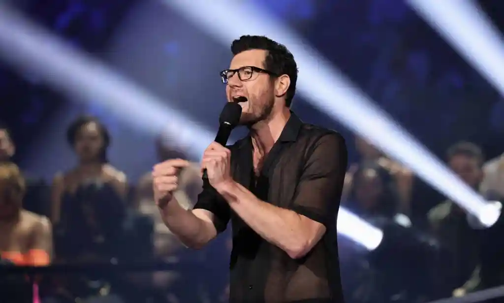 Billy Eichner wears a dark outfit as he gestures towards himself as he speaks at the MTV VMAs