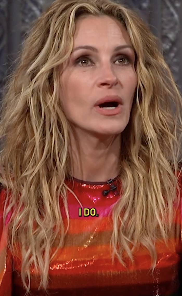 A woman with wavy hair in a shiny, multi-patterned outfit speaks with a focused expression. Subtitles read: "I do."