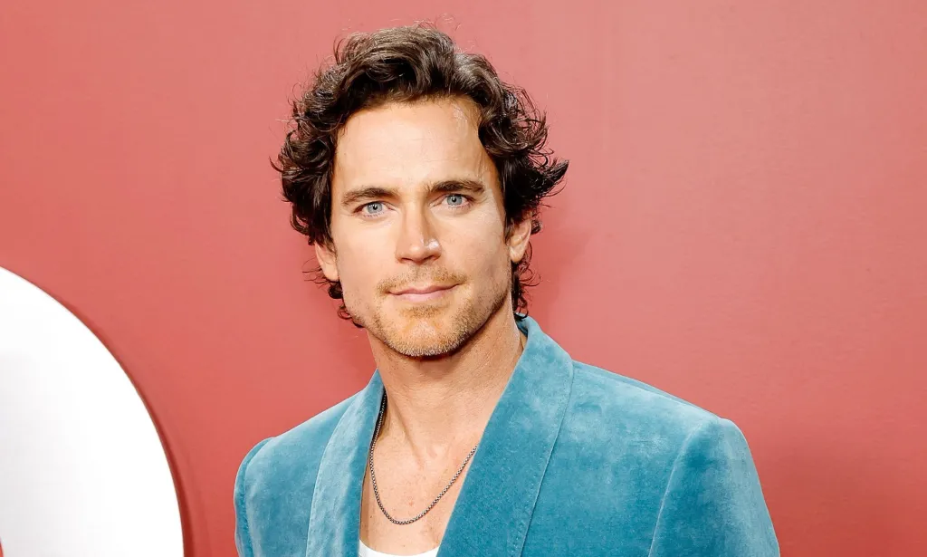 Matt Bomer in a blue blazer and white shirt with silver chain poses for photos against an reddish background.