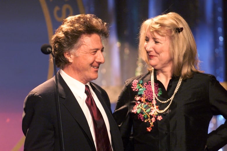 Dustin Hoffman, in a black suit and tie stands, next to a Teri Garr with long blonde hair  and wearing a black blouse with flowers embroidered on her right shoulder. 